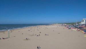 A picture of a beach with many people. A clown image is on a pole in the distance.